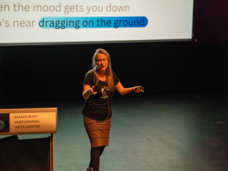 Image of A woman gives a presentation on stage at Mandurah Performing Arts Centre, gesturing with her hands. Behind her, a screen displays text with the words dragging on the ground highlighted in blue.