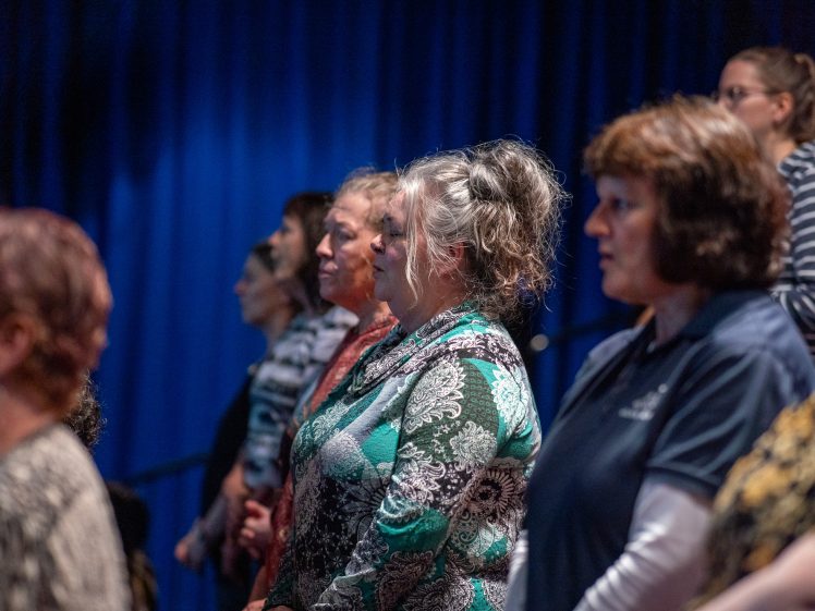 Image of A group of women stand in rows, facing forwards in a dimly lit room with a blue backdrop. The women are of varying ages and wear patterned tops and casual clothing.