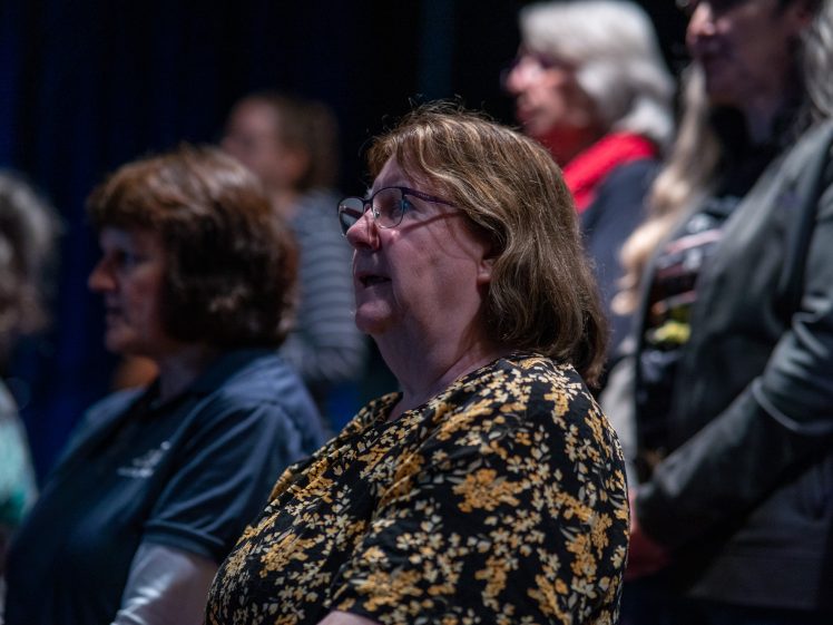 Image of A group of women standing indoors, facing forwards. The focus is on a woman in glasses and a floral blouse, with other women in the background, some slightly blurred. The lighting is dim.