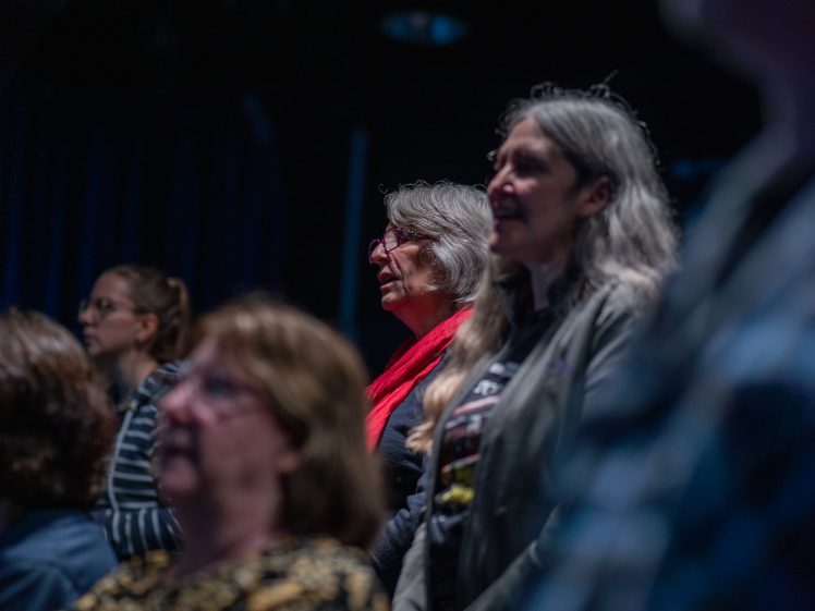 Image of A group of people, mostly women, stand closely together in a dimly lit room, attentively facing forwards. One woman in the centre wears glasses and a bright red scarf.