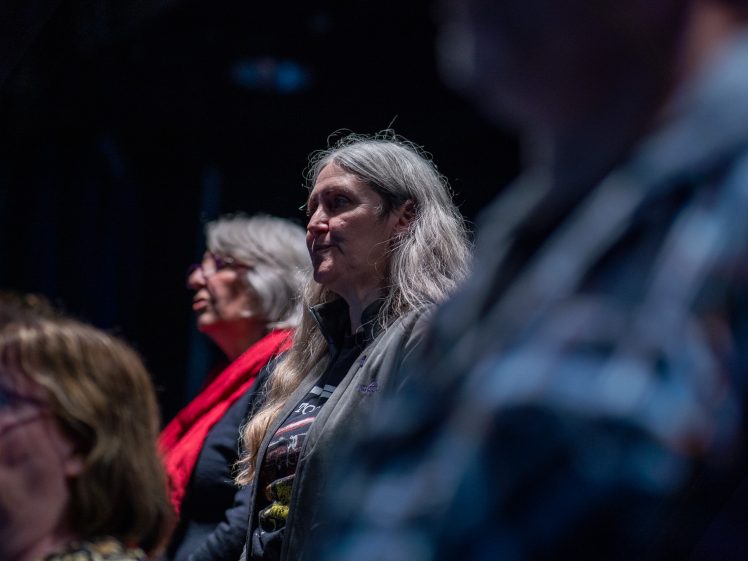 Image of Several people sit or stand in a dimly lit room, facing forwards. The focus is on a person with long grey hair in the centre, and another person with glasses and a red scarf slightly behind.
