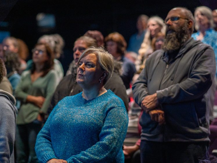 Image of A group of people stands attentively in an indoor setting, appearing focused on something out of view. The woman in the foreground wears a blue jumper, and a man beside her has a grey hoodie and a beard.