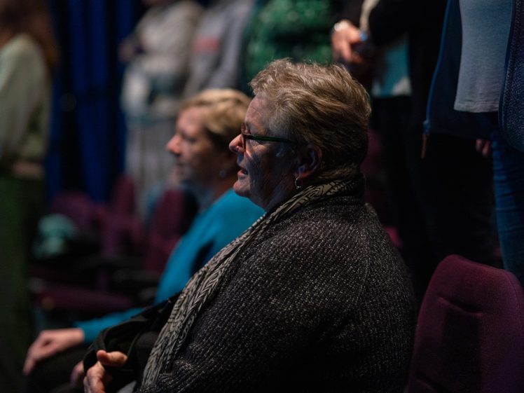 Image of A woman with short grey hair and glasses sits in a dimly lit auditorium, surrounded by others, attentively watching something off-camera. She wears a grey jumper and a patterned scarf.