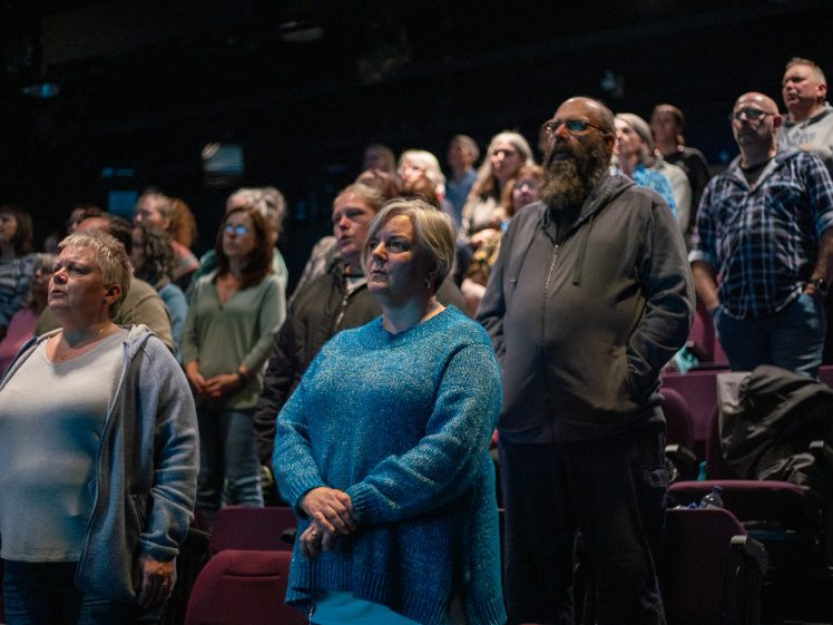 Image of A diverse group of people stands in an auditorium, facing forward with serious expressions. The seats are maroon, and the lighting is dim, suggesting they may be participating in a formal or solemn event.