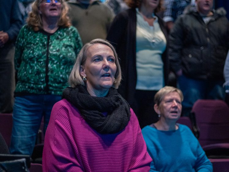 Image of A group of people, mostly women, stand and appear to sing or follow along with something off-camera in a dimly lit auditorium with theatre seating. The focus is on a woman in a pink jumper in the foreground.