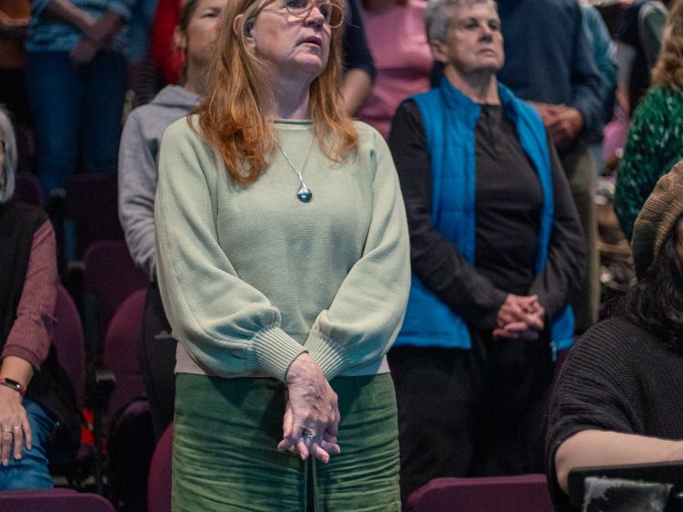 Image of A group of people stand closely together in a dimly lit auditorium, facing forwards. In the foreground, a woman with long red hair, glasses, and green clothing stands with her hands clasped.