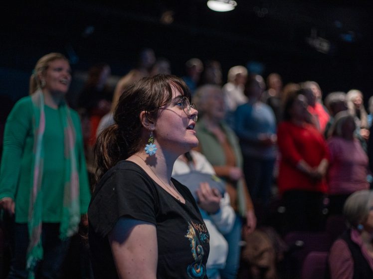 Image of A woman with glasses and earrings stands in the foreground, looking ahead in a dimly lit room, with a group of people behind her, all appearing to watch or listen to something off-camera.