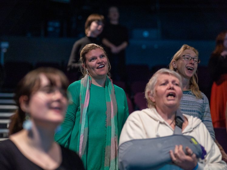 Image of A group of people standing indoors, appearing to sing or speak together. The foreground shows three women, one holding a cushion, all engaged and expressive, with others visible in the background.