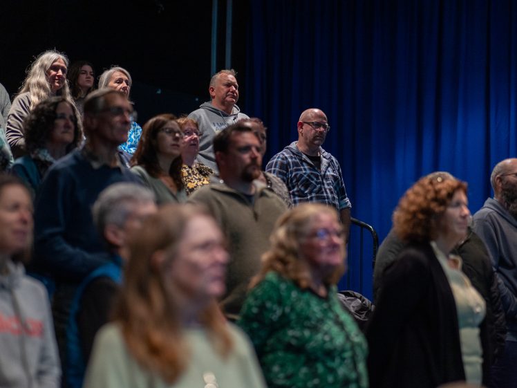 Image of A group of people of various ages and genders stand closely together in a dimly lit room with blue curtains in the background, looking forward with attentive expressions.