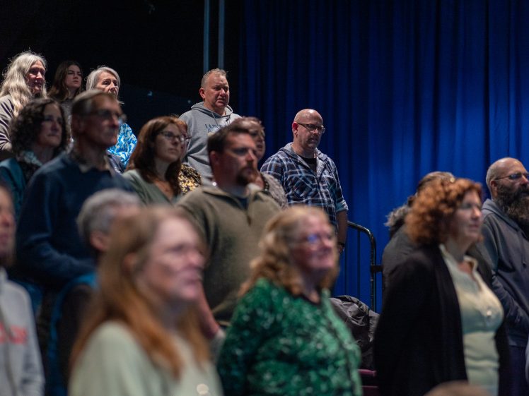 Image of A group of adults stands in an auditorium, facing forwards. The background features a blue curtain, and the audience members appear attentive and focused.