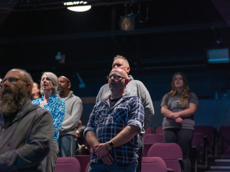 Image of A group of people stand in a dimly lit auditorium, facing forward with serious expressions; empty purple seats are visible around them.