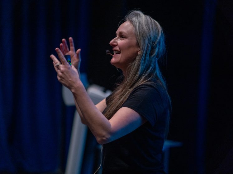 Image of A woman with long grey hair, wearing a black shirt and a headset microphone, gestures enthusiastically whilst speaking on stage against a dark background.