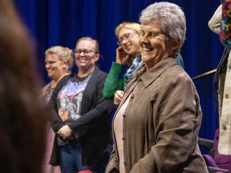 Image of A group of women, including an older woman in the foreground smiling, stand together in a room with purple chairs and a dark blue curtain in the background.