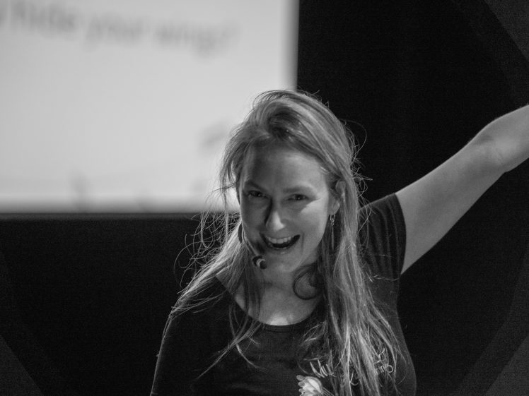 Image of A woman with long hair smiles enthusiastically, wearing a headset microphone and raising one arm. She stands in front of a blurred screen in a black-and-white setting, suggesting a presentation or performance.