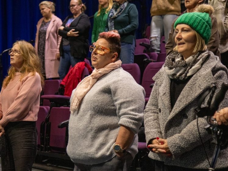 Image of A group of people standing in theatre-style seats, appearing to sing or speak. Three women in the front row wear winter clothing, including jumpers, scarves, and a green knitted hat.