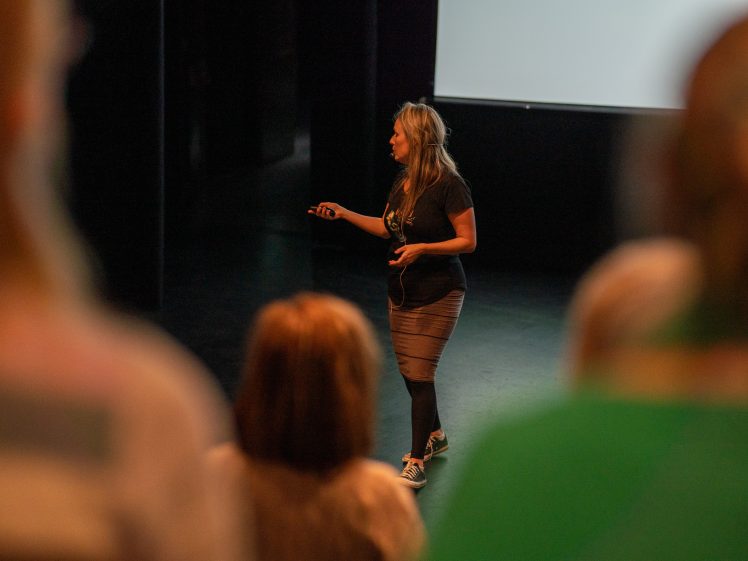 Image of A woman stands and speaks on stage in front of an audience, holding a remote, with a blank screen behind her. The image is taken from the perspective of the seated audience.