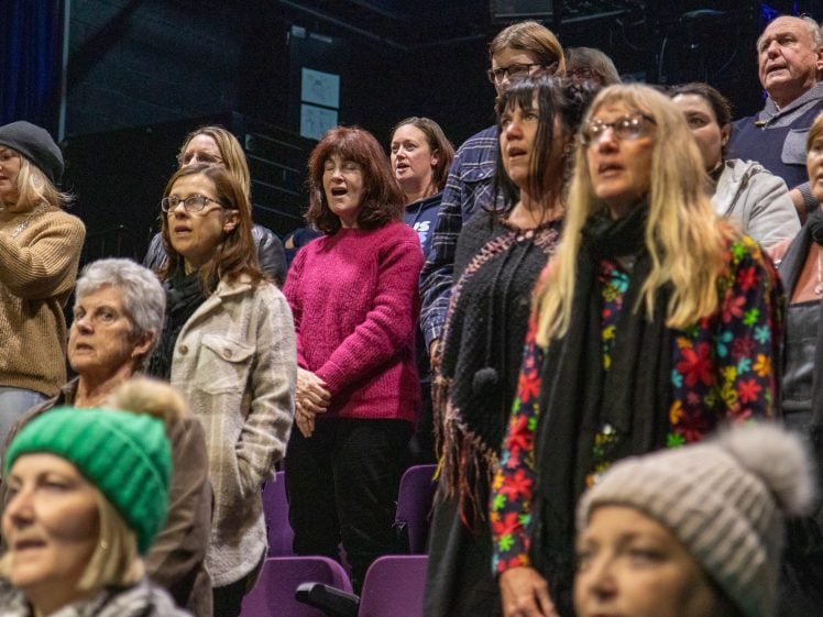 Image of A group of people of various ages stand in rows, singing together indoors. Some wear winter clothing like hats and scarves, and the background is dimly lit, suggesting a rehearsal or choir practice.