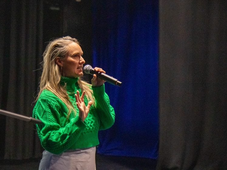 Image of A person with long blond hair, wearing a bright green jumper, speaks into a microphone on a dimly lit stage with dark curtains and blue lighting in the background.