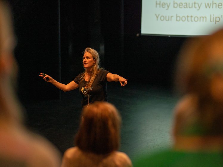 Image of A woman stands on a stage, gesturing with both arms extended, speaking to an audience. Part of a projected screen with partial text is visible behind her, and several blurred figures are in the foreground.