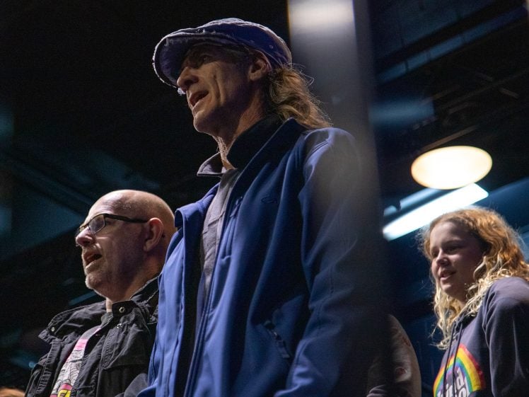 Image of Three people stand indoors under artificial lighting, looking ahead. One wears glasses and a black jacket, the second has long hair, a blue jacket, and a cap, and the third has wavy hair and a dark hoodie with a colourful design.