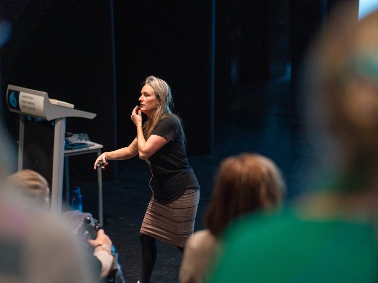 Image of A woman stands near a lectern, speaking to an audience in a dimly lit auditorium. She holds a microphone and gestures thoughtfully, engaging with the crowd. Several people are visible in the foreground, listening attentively.