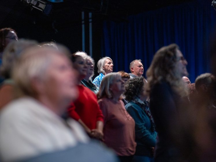 Image of A group of people stand closely together in a dimly lit room with blue curtains, all facing the same direction and appearing attentive or focused.