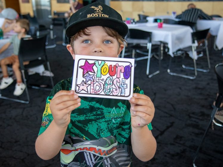 Image of A young child wearing a Boston cap holds up a colourful hand-drawn sign that reads You’re the Best with stars and hearts, smiling in a room with tables and people in the background.
