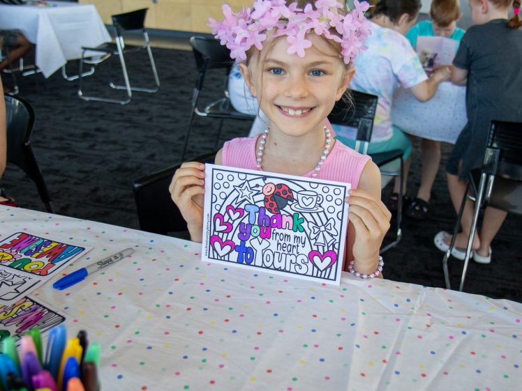 Image of A smiling child wearing a pink flower crown holds up a colourful Thank You from my heart to yours card at a table with felt-tip pens and colouring sheets. Other children and adults are seated in the background.