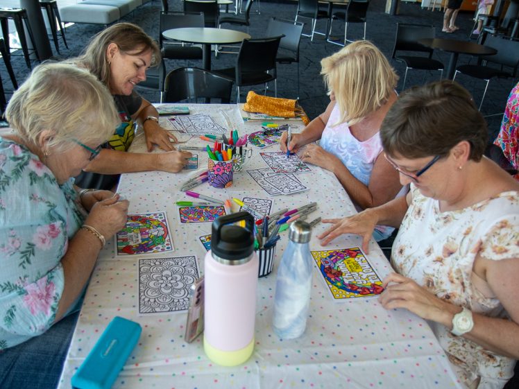 Image of Four women sit around a table covered with felt-tip pens and colouring pages, each focused on colouring intricate designs. The scene is relaxed and sociable in a well-lit indoor space with tables and chairs in the background.