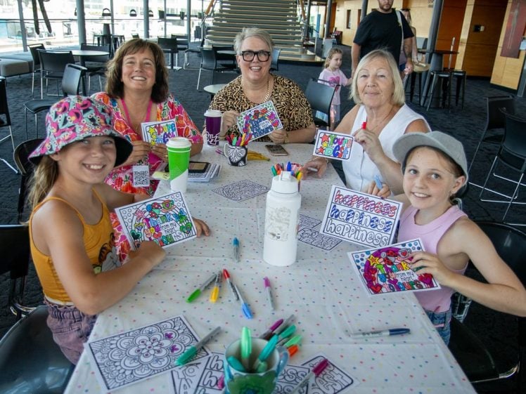 Image of Five smiling people, including two girls and three women, sit around a table displaying colourful art cards they have made. The table is covered with art supplies, and a child stands in the background.