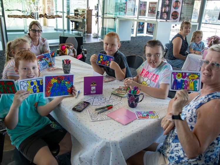 Image of A group of smiling children and adults sit around a table covered with art supplies, proudly holding up colourful, hand-decorated cards with positive messages like “YOU ARE AWESOME.” They appear to be in a bright, modern room.