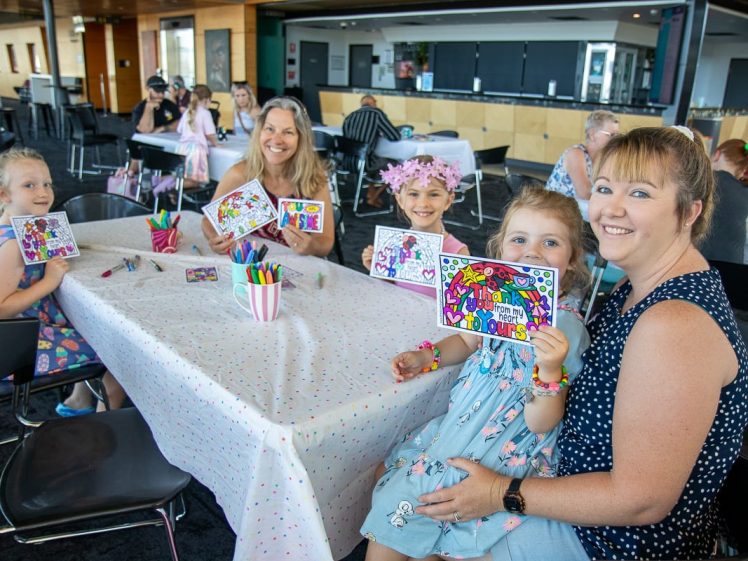 Image of A group of smiling children and adults sit at a decorated table, proudly holding up colourful birthday cards they’ve made with felt-tip pens, in a bright, cheerful indoor setting.