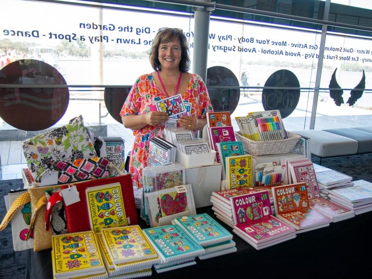 Image of A smiling woman stands behind a table filled with colourful journals, colouring books, pencils, and art supplies, displayed in a bright indoor space with large windows in the background.