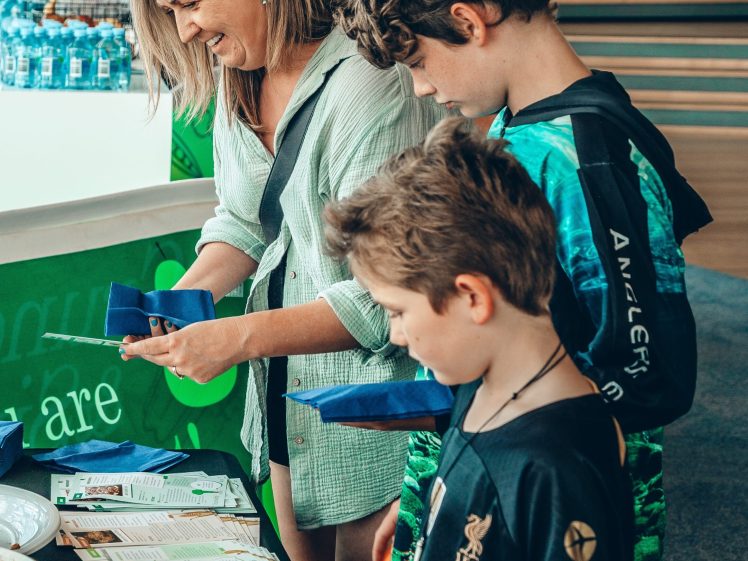 Image of A woman and two boys stand at a table with leaflets, bottles, and other items, engaging with the display. The background shows water bottles and people in a bright, indoor setting.