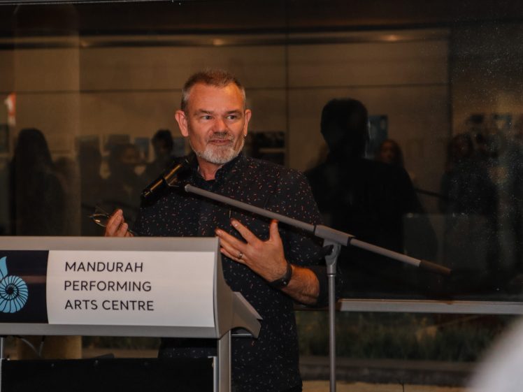 Image of A man with a beard speaks at a podium labelled Mandurah Performing Arts Centre, gesturing with one hand, with a microphone and blurred audience in the background.