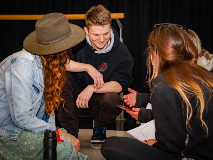 Image of Three young adults sit on the floor in a circle, talking and smiling. One person in a hat faces away, another holds a mobile, and a man in the middle looks at them, appearing engaged and happy. Black curtain in the background.