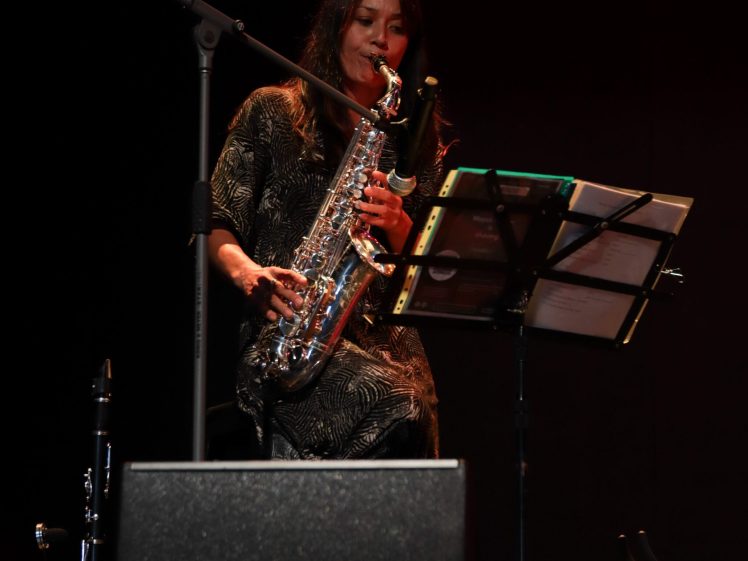 Image of A woman sits on stage playing a saxophone, with sheet music in front of her on a stand and a microphone positioned nearby. The background is dark, focusing attention on the musician.