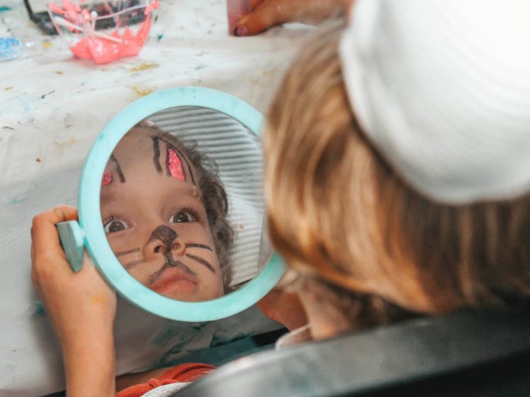 Image of A child with face paint resembling a cat looks into a round hand mirror, reflecting their face with painted whiskers and a nose at a craft-covered table.