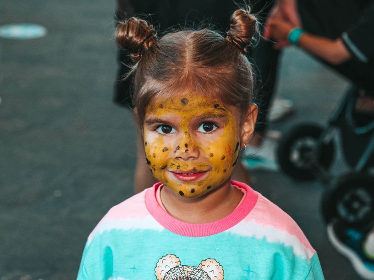 Image of A young girl with brown hair in two buns has her face painted like a yellow leopard. She is wearing a colourful sweatshirt with a bear design and is smiling at the camera. The background is blurred with people and a pushchair.