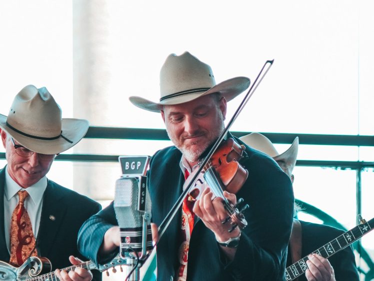 Image of Three men in suits and cowboy hats play string instruments and perform near a vintage microphone. The central man smiles while playing the violin, and the background is softly lit with natural light.