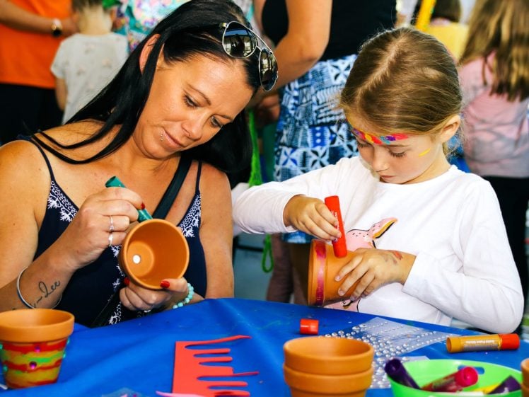 Image of A woman and a young girl sit at a table, painting terracotta pots together. The table is covered with art supplies and decorations. The girl has face paint and is focused on her pot, whilst the woman carefully paints hers.