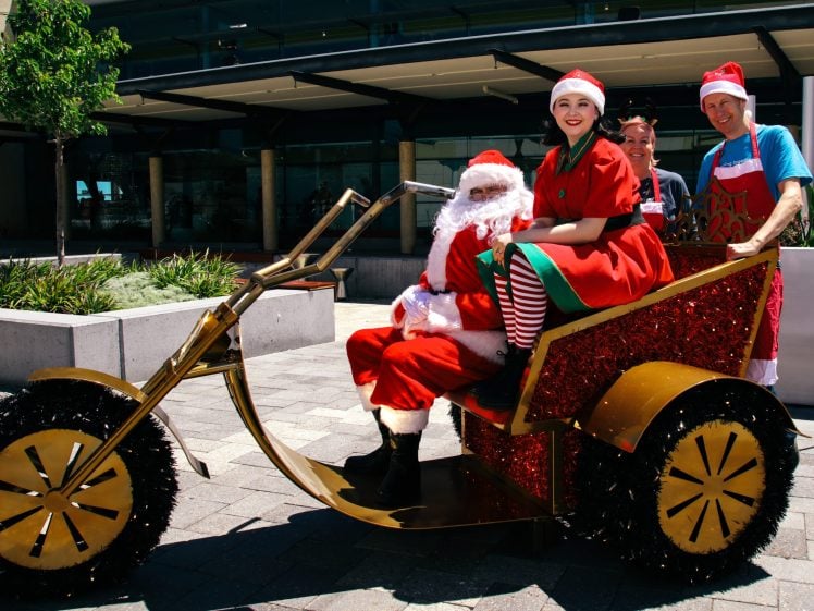Image of Four people in festive costumes, including Father Christmas and two elves, sit on a decorated tricycle with sparkly wheels outdoors on a sunny day, smiling at the camera.