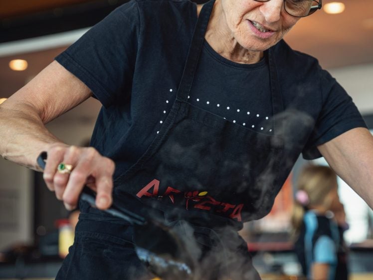 Image of An older person wearing glasses and a black apron is stirring or serving food from a steaming pot filled with orange vegetables in a kitchen setting.
