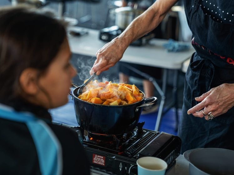 Image of A person stirs a pot of steaming vegetables on a portable gas cooker, while another person in the foreground watches attentively. The setting appears to be a casual, indoor cooking environment.