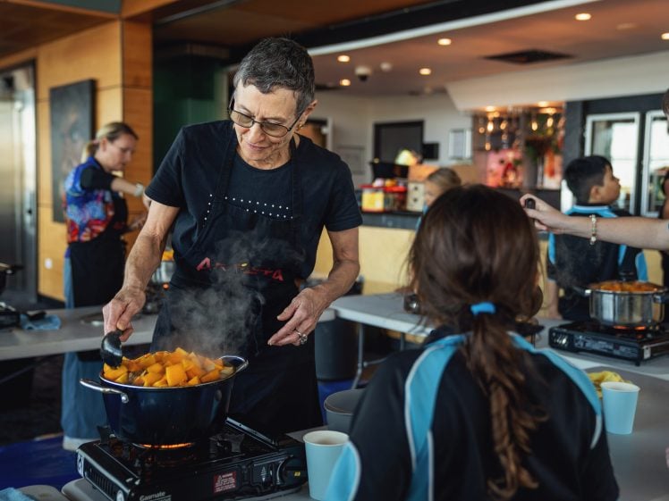 Image of An older woman stirs a pot of steaming food containing chopped vegetables whilst teaching children in a kitchen classroom setting. Other people and cooking supplies are visible in the background.
