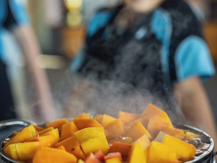 Image of A pot filled with steaming, chopped orange and yellow vegetables sits in the foreground, whilst a smiling child wearing a blue shirt and dark apron appears blurred in the background.