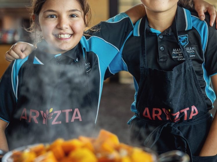 Image of Two smiling children in matching blue uniforms and black aprons stand side by side with arms round each other, behind a steaming pot filled with orange vegetables in a kitchen or canteen setting.