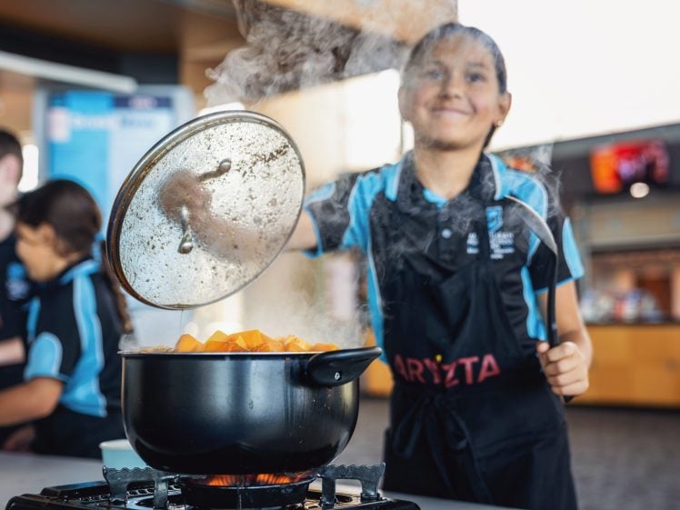 Image of A smiling child lifts the lid off a steaming saucepan filled with chopped vegetables while cooking in a kitchen, wearing a dark apron and blue shirt. Other children are visible in the background.