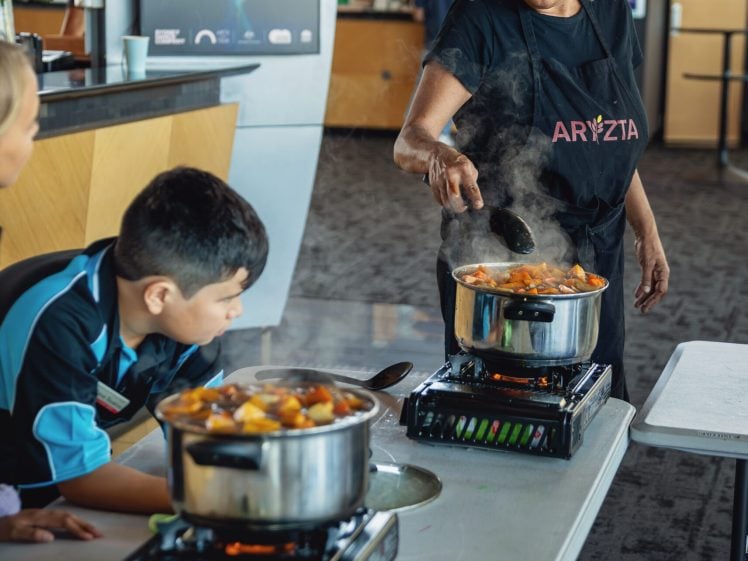 Image of A woman wearing a black apron stirs food in a pot on a portable hob, with a young boy watching beside her. Both are at a table in an indoor setting, with steam rising from the pots.