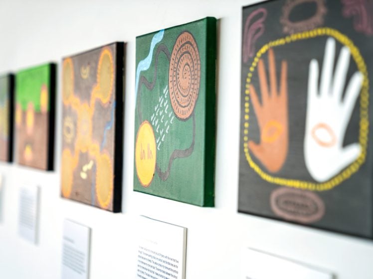Image of A row of colourful, abstract paintings with Indigenous-style patterns and hand motifs is displayed on a white wall, each accompanied by a small descriptive card below.